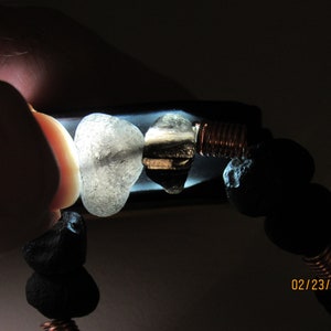 May include: A bracelet featuring black lava rock beads, clear and dark stones, and copper wire accents. The bracelet is held up to a light source, highlighting the textures and colors of the stones. The date 02/23/2021 is visible.