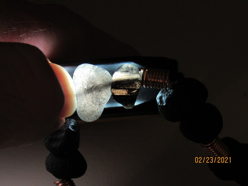 May include: A bracelet featuring black lava rock beads, clear and dark stones, and copper wire accents. The bracelet is held up to a light source, highlighting the textures and colors of the stones. The date 02/23/2021 is visible.