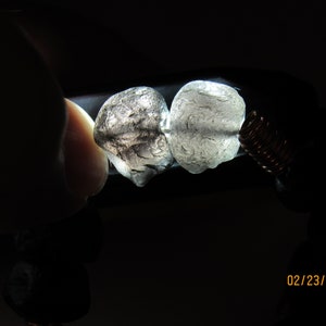 May include: Two translucent, irregularly shaped stones are held in place by a copper wire coil, with a dark background. The stones are light gray and have a textured surface. The bracelet also features dark, round beads.