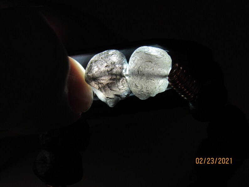 May include: Two translucent, irregularly shaped stones are held in place by a copper wire coil, with a dark background. The stones are light gray and have a textured surface. The bracelet also features dark, round beads.