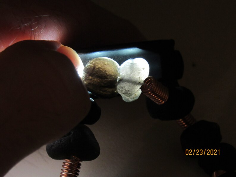 May include: A handmade bracelet featuring black, rounded beads, and two lighter-colored, irregularly shaped stones. The bracelet incorporates copper wire accents, with a date stamp of 02/23/2021 visible in the image.