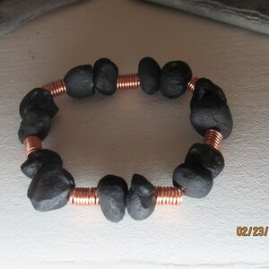 May include: A handmade bracelet featuring dark gray, irregularly shaped stones strung together with copper wire. The bracelet is circular and appears to be made of natural materials. The copper wire is coiled between the stones.