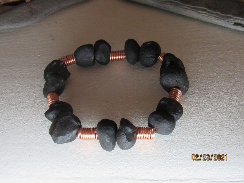 May include: A handmade bracelet featuring dark gray, irregularly shaped stones strung together with copper wire. The bracelet is circular and appears to be made of natural materials. The copper wire is coiled between the stones.