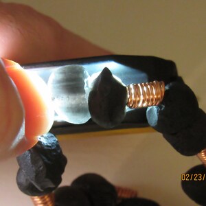 May include: Close-up of a bracelet featuring dark, irregularly shaped beads strung together with copper wire. The beads are a deep black color, and the wire has a warm, reddish-brown hue. The bracelet is illuminated by a bright light.
