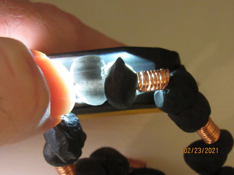 May include: Close-up of a bracelet featuring dark, irregularly shaped beads strung together with copper wire. The beads are a deep black color, and the wire has a warm, reddish-brown hue. The bracelet is illuminated by a bright light.