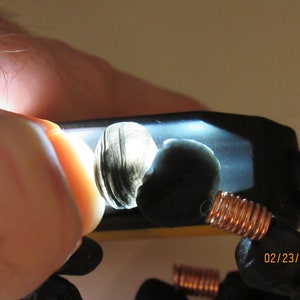 May include: Close-up of two dark, polished stones illuminated by a bright light. One stone has a layered, shell-like appearance. Copper wire coils are visible, suggesting a jewelry component. The date 02/23/2021 is in the lower right corner.