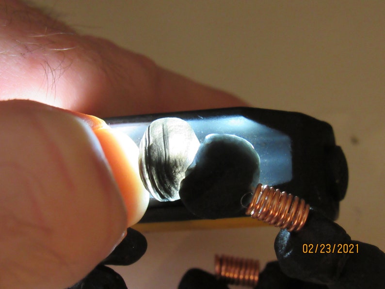 May include: Close-up of two dark, polished stones illuminated by a bright light. One stone has a layered, shell-like appearance. Copper wire coils are visible, suggesting a jewelry component. The date 02/23/2021 is in the lower right corner.