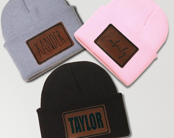 May include: Three knitted beanies with faux leather patches. The beanies are gray, pink, and black. The patches have the names "Xander", "Emberly", and "Taylor" written on them.