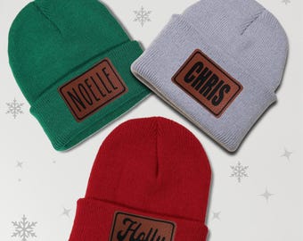 May include: Three knitted beanies in green, gray, and red with brown leather patches. The patches have the names "Noelle", "Chris", and "Holly" written in black.