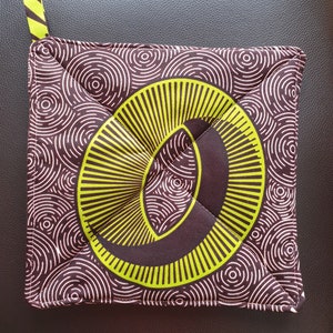 May include: Square, quilted oven mitt with a brown and white circular pattern. The centre features a large, stylized letter "O" in bright yellow-green. A small loop of the same yellow-green fabric is attached to one corner.
