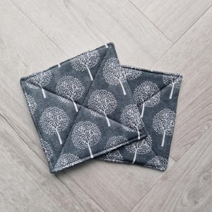 May include: Two square, quilted pot holders with a grey background and a white tree pattern. The pot holders are made of fabric and have a stitched, cross-like design in the centre.