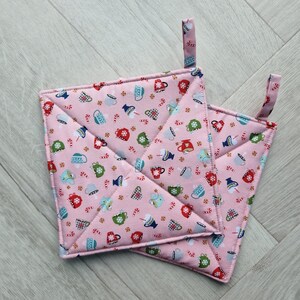 May include: Two pink quilted pot holders with a repeating pattern of mugs, snowflakes, and candy canes. Each square pot holder has a loop for hanging. The pot holders are made of fabric and have a stitched edge.
