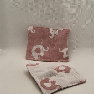 May include: Two square fabric coasters. The coasters are a dusty rose colour with a white elephant pattern. The coasters are made of a textured fabric. One coaster is in the foreground, and the other is in the background.