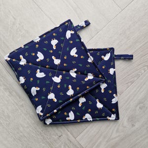 May include: Two navy blue, quilted pot holders with a white goose and floral pattern. Each square pot holder has a small fabric loop for hanging. The pot holders are approximately 20 cm square.
