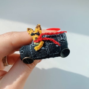 May include: A small, yellow, crocheted dog wearing sunglasses and a red scarf is driving a black, crocheted car with a button for a wheel.