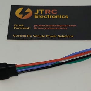 Puede incluir: Un conector negro con múltiples cables de colores, incluyendo rojo, azul y verde. En el fondo, una tarjeta de visita con el texto "JTRC Electronics" e información de contacto. La tarjeta también indica "Custom RC Vehicle Power Solutions."