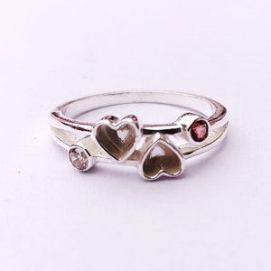 May include: A silver ring featuring three heart-shaped designs. The ring has a small pink stone and a clear stone.