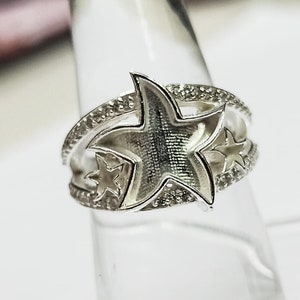 Pode incluir: Anel de prata com três desenhos em forma de estrela. A estrela central é maior e tem uma superfície texturizada. O anel é adornado com pequenas pedras transparentes.