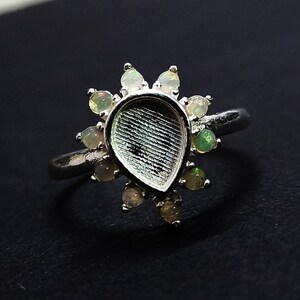 May include: A silver ring with a teardrop-shaped setting surrounded by small, clear stones. The ring is designed to hold a larger stone in the centre.