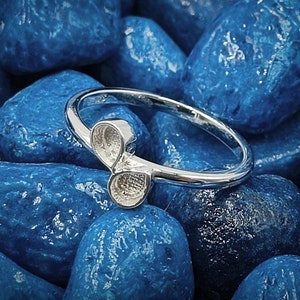 Puede incluir: Un anillo de plata con un diseño único de trébol de tres hojas. El anillo está fotografiado sobre un lecho de piedras azules.