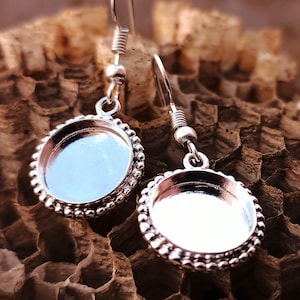 Könnte beinhalten: Ein Paar silberne Ohrringe mit runden, gefassten, leeren, silberfarbenen, runden Cabochons. Die Ohrringe haben einen perlenbesetzten Rand und einen silbernen Haken.