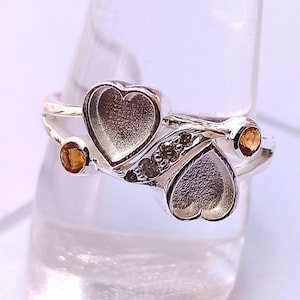 May include: Silver ring with two heart-shaped settings and small orange gemstones. The ring is decorated with a row of smaller gemstones.
