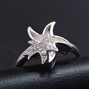 Pode incluir: Um anel de prata com um design de estrela do mar. A estrela do mar é adornada com pequenas pedras transparentes.