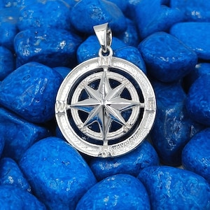 May include: A silver pendant with a compass rose design. The pendant has a star-shaped design in the centre, surrounded by a circular frame. The pendant is set against a background of blue stones.