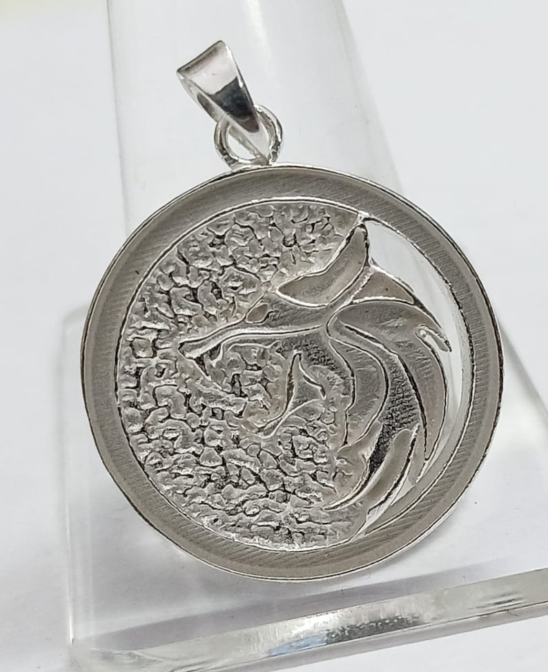 May include: Silver pendant featuring a detailed, textured design of a dragon with its wings spread. The pendant is round and has a smooth, polished edge.