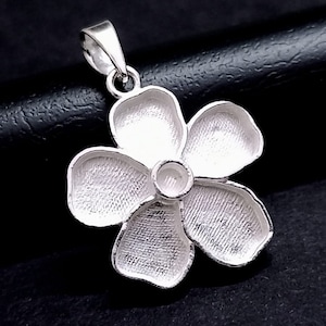 May include: A silver flower pendant with five petals and a central circular detail. The pendant has a textured surface and a loop for a chain. The background is black.