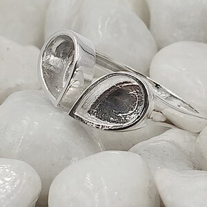 May include: A silver ring with two teardrop-shaped settings. The ring is set against a background of white stones. The settings are empty, ready for gemstones or other embellishments. The ring's design is modern and minimalist.