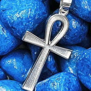Könnte beinhalten: Ein silberfarbener Ankh-Anhänger, ein Symbol für ewiges Leben, liegt vor einem Hintergrund aus leuchtend blauen Steinen. Das Design des Anhängers zeigt eine Schlaufe oben und eine Kreuzform unten mit strukturierten Details.