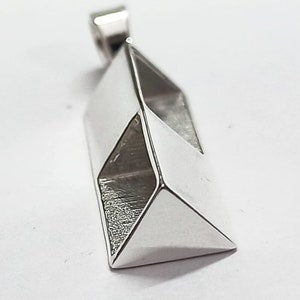May include: A silver pendant with a geometric design. The pendant has a rectangular shape with angled facets, creating a three-dimensional effect. The top features a small loop for a chain. The metal has a polished, reflective surface.