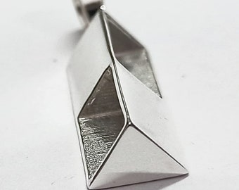 Op de afbeelding: Een zilveren hanger met een geometrisch ontwerp. De hanger heeft een rechthoekige vorm met schuine facetten, waardoor een driedimensionaal effect ontstaat. De bovenkant heeft een kleine lus voor een ketting. Het metaal heeft een gepolijst, reflecterend oppervlak.