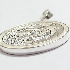 May include: Silver pendant with a detailed, textured design of a stylized wave. The pendant is oval shaped and has a smooth, polished finish.