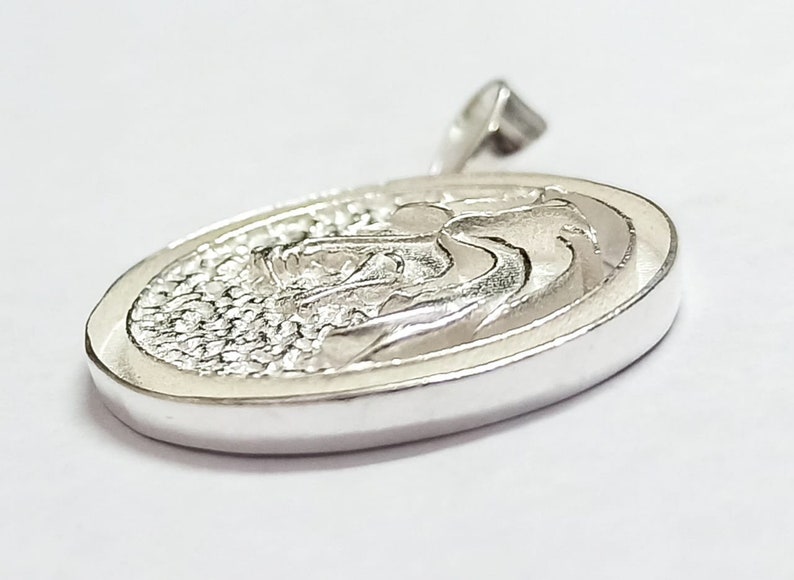 May include: Silver pendant with a detailed, textured design of a stylized wave. The pendant is oval shaped and has a smooth, polished finish.