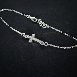 May include: A silver bracelet with a small cross pendant. The cross is the focal point of a fine chain, secured with a clasp. The bracelet is set against a dark backdrop, accentuating its reflective quality.