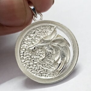 May include: Silver pendant featuring a detailed dragon design with a textured background. The pendant is circular and has a loop at the top for attaching a chain.