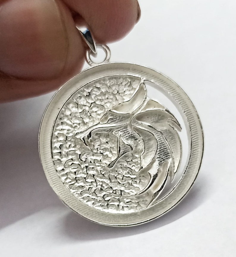May include: Silver pendant featuring a detailed dragon design with a textured background. The pendant is circular and has a loop at the top for attaching a chain.