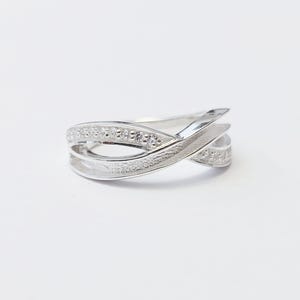 May include: A silver ring with an unusual, interwoven design. The ring has a band with small, sparkling stones and a flowing, layered structure. The ring is set against a white background, highlighting its elegant form.