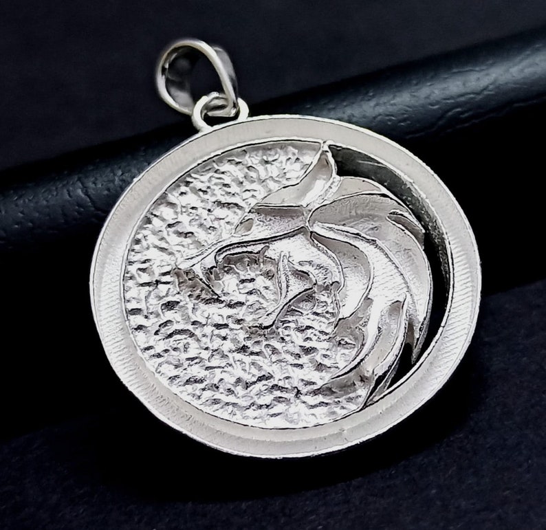 May include: A silver pendant featuring a detailed wolf head design. The pendant is round and has a textured surface. The wolf's head is facing to the right and has a sharp, detailed design.