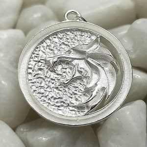 May include: A silver pendant featuring a detailed, textured design of a wolf's head with a circular frame.