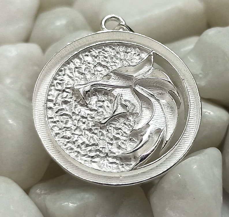 May include: A silver pendant featuring a detailed, textured design of a wolf's head with a circular frame.