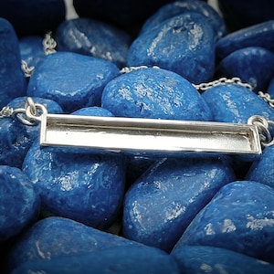 May include: A silver bar pendant necklace with a chain. The pendant is rectangular and has a smooth, polished finish. The necklace is displayed on a bed of blue stones.