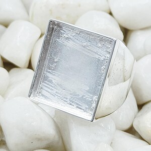 May include: A silver ring with a square-shaped white stone set in the center. The ring is on a bed of white stones.