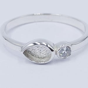 Op de afbeelding: Een zilveren ring met een traanvormige zetting en een kleinere ronde steen. De ring is simpel en elegant.