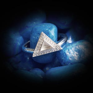Può includere: Un anello in argento con un elemento centrale triangolare caratterizzato da un bordo pavé di piccole pietre chiare che circonda un centro metallico triangolare liscio e incassato. Il testo Thevelvetsilver è visibile sulla fascia dell'anello.