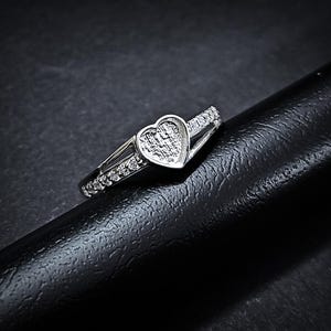 Könnte beinhalten: Silberner Ring mit herzförmigem Mittelstein und kleinen, klaren Akzentsteinen. Der Ring hat ein geteiltes Banddesign. Das Herz ist strukturiert.