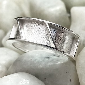 May include: A silver ring with a geometric design. The ring has a textured surface and a wide band.