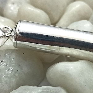May include: A silver, cylindrical pendant with a screw-on top. The pendant is lying on a bed of white stones.